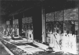 unscharfe Schwarz-Weiß-Fotografie, rechts vier Öfen mit jeweils vier Öffnungen, links vor den Öfen Schienen