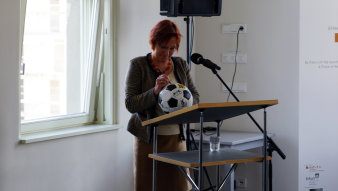 Farbfoto; eine Frau steht am Rednerpult und unterschreibt auf einem Fußball