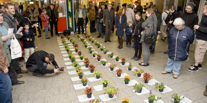 Bahnhofshalle, in der Bildmitte zwei in Zweierreihen aufgestellte Blumentöpfe mit Blumen in unterschiedlichen Farben,jeder Blumentopf steht auf einem ca. A-4 großen Blatt Papier, um die Blumentöpfe herum eine größere Menschenansammlung