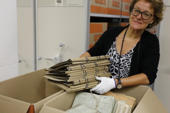 Foto: Stadtarchiv Aktenfund © Stadtverwaltung Erfurt Dr. Antje Bauer hält einen Stapel Akten in der Hand, vor ihr stehen zwei mit Unterlagen gefüllte Kartons, im Hindergrund sieht man ein Regal mit vielen Kartons.