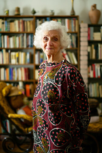 Eine ältere Frau steht, gekleidet in ein buntes Kleid, in einer Wohnung. Im Hintergrund sind Bücherregale zu sehen.