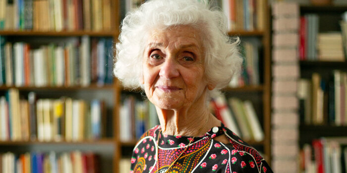 Eine ältere Frau steht, gekleidet in ein buntes Kleid, in einer Wohnung. Im Hintergrund sind Bücherregale zu sehen.