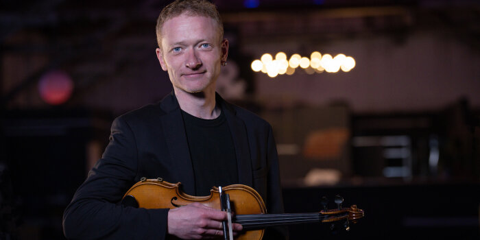 ein Mann mit Violine unter dem Arm steht in einem dezent beleuchteten Raum