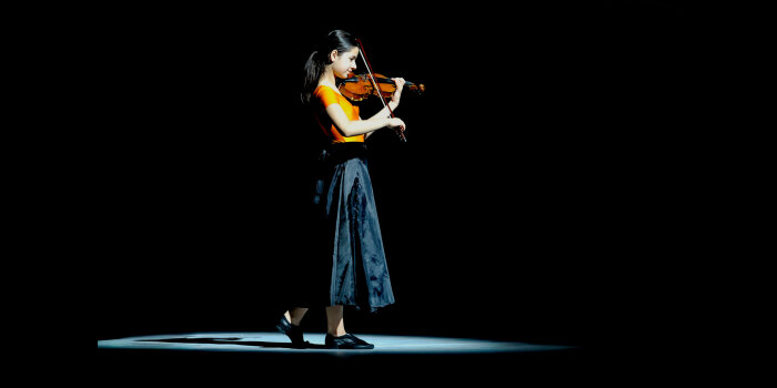 Eine junge Frau mit langen Haaren steht auf einer Bühne und spielt Violine.