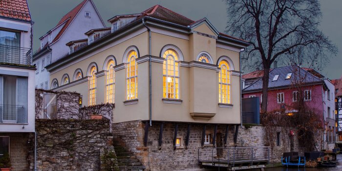 Foto: Kleine Synagoge, Erfurt © Stadtverwaltung Erfurt das gelbe Gebäude, mit hell erleuchteten Fenstern unterm Nachthimmel vom Wasser aus fotografiert