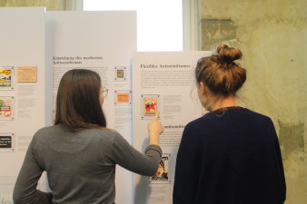 Zwei Frauen stehen vor Ausstellungstafeln. Eine der Frauen zeigt auf einen Sticker auf der Tafel. 