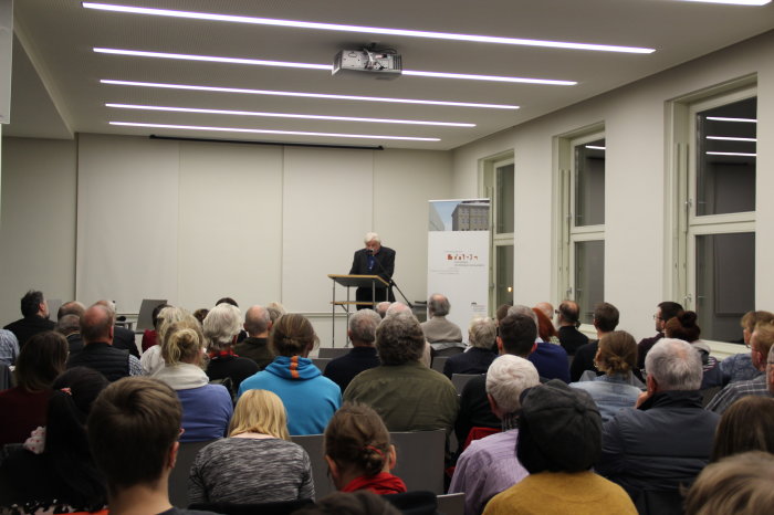 Prof. Dr. Wolfgang Benz bei seinem Vortrag im Erinnerungsort Ein Mann steht hinter einem Rednerpult und spricht in ein Mikrofon.
