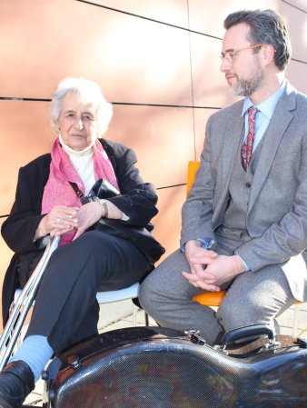 Eine ältere Frau und ein jüngerer Mann sitzen nebeneinander auf zwei Stühlen. Vor ihnen liegt ein Cellokoffer.