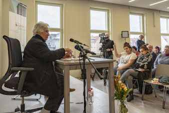 Eine ältere Frau spricht an einem Tisch sitzend in ein Mikrofon. 