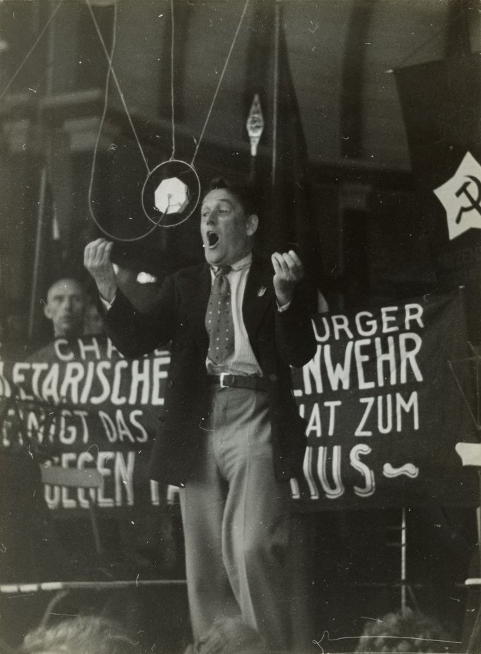 Ein Mann steht auf einer Bühne. Er gestikuliert und spricht animiert. Hinter ihm ist ein Banner angebracht.
