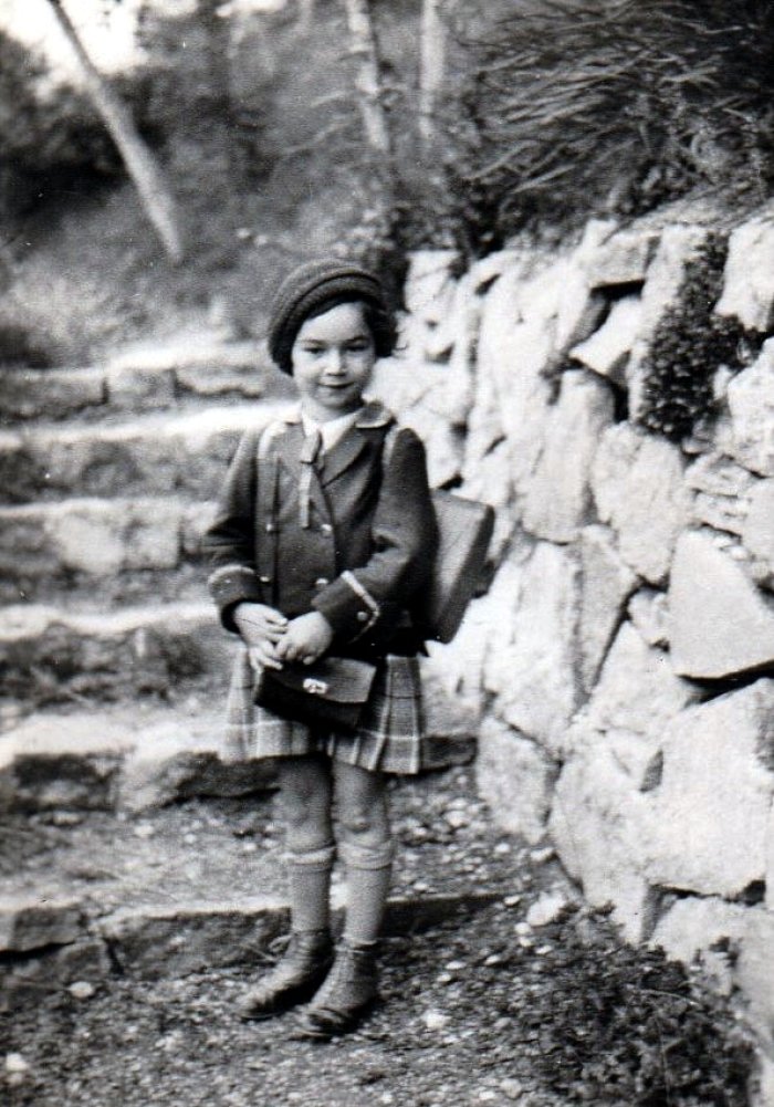 Ein junges Mädchen, mit Mütze, Jacke, Rock, Schulranzen. Es handelt sich um ein älteres schwarz-weiß Bild. 