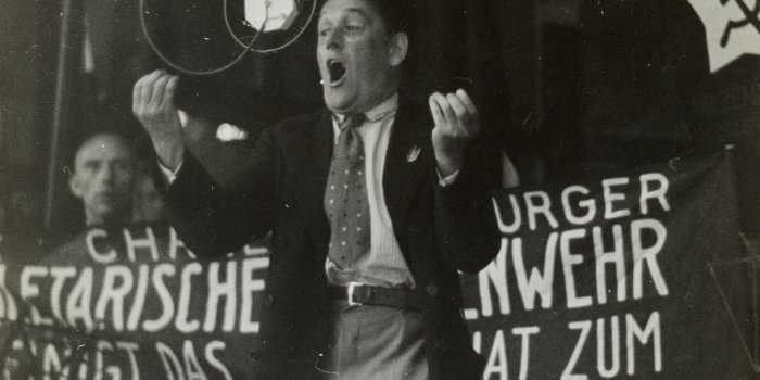 Ein Mann spricht enthusiastisch in ein hängendes Mikrofon. Hinter ihm wird ein Banner hochgehalten. 