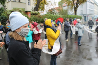 Über zwanzig Menschen stehen in Winterkleidung, mit Masken auf einem Platz und klatschen. 