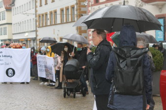 Menschen mit Transparenten stehen im Regen auf einem Platz, ein Mann spricht ins Mikrofon.