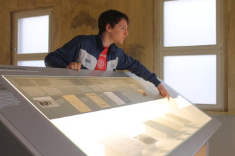 Ein junger Mann steht an einem Ausstellungspult und deutet auf ein Dokument. 