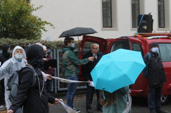 Ein älterer Mann spricht in ein Mikro, um ihn herum Menschen mit Regenschirmen.
