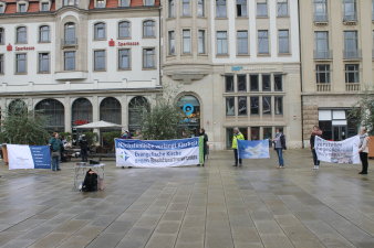 Mehrere Menschen stehen mit Transparenten auf einem Platz, im Hintergrund Stadthäuser. 