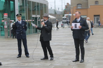 Drei Männer: Der linke mit Polizeiuniform, der mittlere spricht in ein Mikro, der rechte hält ein Bild hoch.