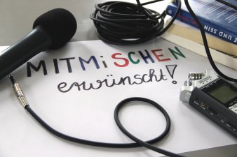 Mikrofone, Kabel, drei Bücher liegen drapiert um ein Blatt Papier auf dem „Mitmischen erwünscht“ steht.