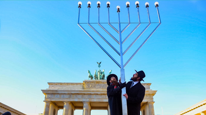 Zwei Männer stehen vor dem Brandenburger Tor. Ihre Hände stützen einen neun-armigen Leuchter. 