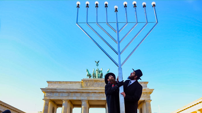 Jüdisches Leben in Berlin: Zur Eröffnung des Jüdischen Bildungszentrums Chabad lässt Rabbi Yehuda Teichtal einen riesigen Chanukka-Leuchter vor dem Brandenburger Tor aufstellen. Zwei Männer stehen vor dem Brandenburger Tor. Ihre Hände stützen einen neun-armigen Leuchter.