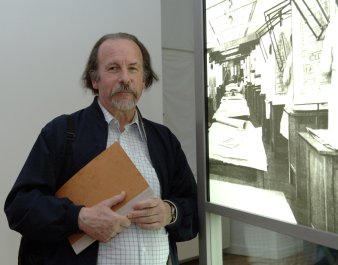 Ein älterer Mann mit Bart steht vor einer Ausstellungstafel. In den Händen hält er eine Mappe.