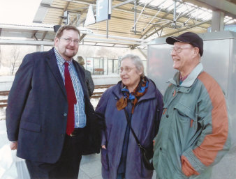 Eine ältere Frau steht zwischen zwei Männern auf einem Bahnsteig. Alle lächeln. 
