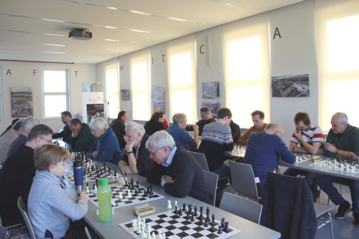 Farboto. Mehrere Personen sitzen an zwei langen Tischen und spielen Schach.