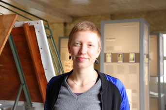 Ida Forbriger, Trainee im Projekt „Mit Erinnerung Demokratie und Vielfalt stärken“ Eine Frau mit grauem Oberteil in einer Ausstellung.