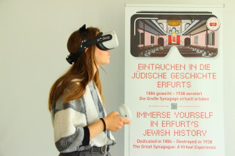 junge Frau mit VR Brille auf dem Kopf daneben ein Plakat mit der Aufschrift Eintauchen in die jüdische Geschichte  