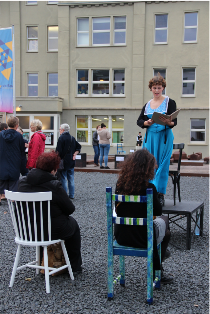 junge Frau auf Posest mit einem blauen Kleid die aus einem Buch vorliest, davor sitzen und stehen Menschen
