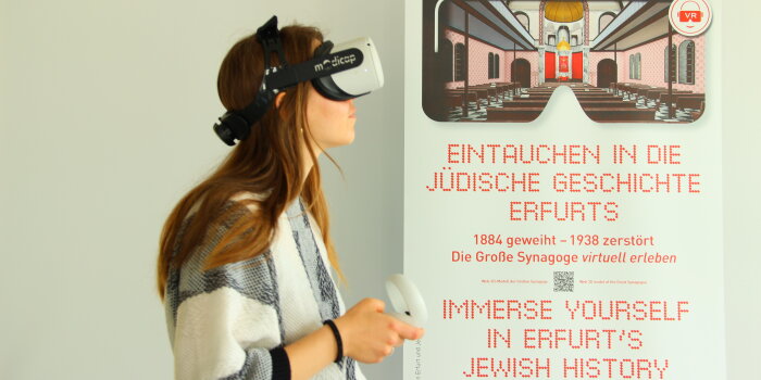 junge Frau mit VR Brille auf dem Kopf daneben ein Plakat mit der Aufschrift Eintauchen in die jüdische Geschichte  