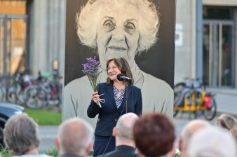 Frau am mikrofon mit Blumrn in der Hand, im Hintergurun ein schwarzweiß Bild einer älteren Frau
