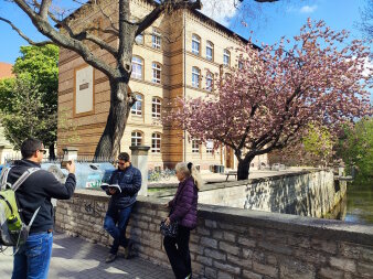 Ein Mann macht ein Foto von einem Mann, der an einer Mauer lehnt und ein Buch liest, nebenan steht eine Frau