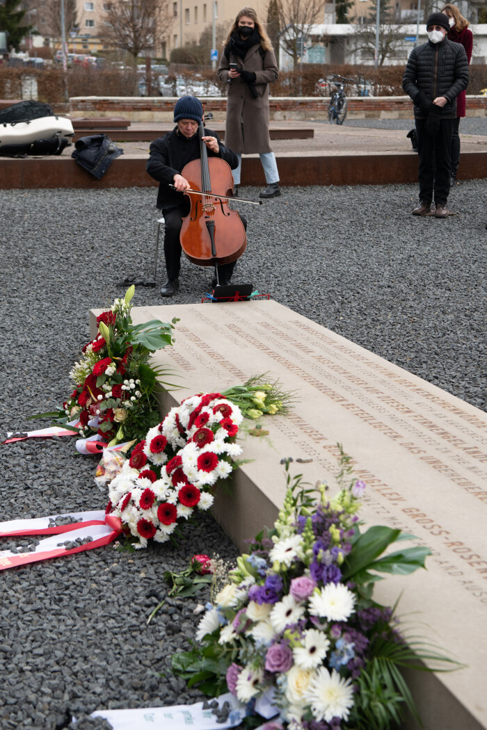 Person mit Cello, an einem Gedenkstein mit Blumen