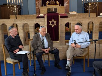 Drei Männer sitzen in einer Synagoge vor der Bima (jüdischer Altar) und führen ein Podiumsgespräch.