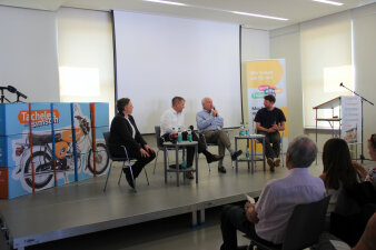Vier Personen sitzen auf einem Podium mit Mikros und sprechen zu einem Publikum.