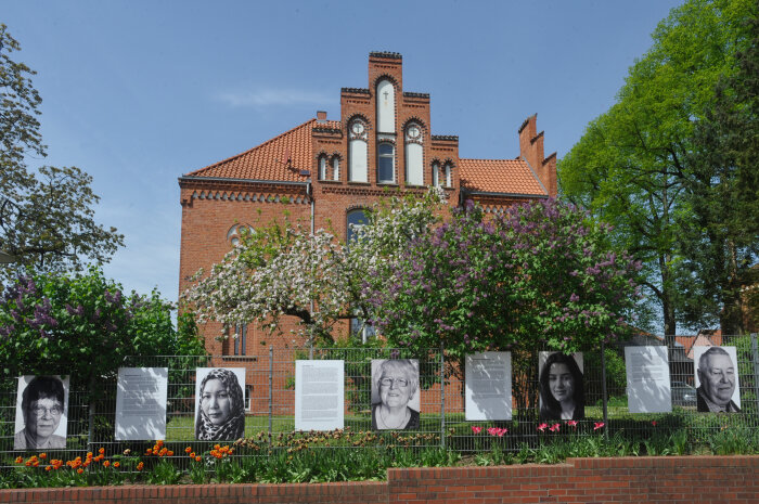 Im Hintergrund ein großes Backsteinhaus mit Garten und im Vordergrund ein Zaun mit angehängten Portraits und Texten