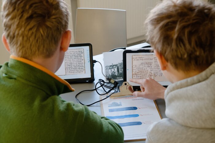 Zwei Schüler sitzen vor einem Arbeitsblatt mit zwei Tablets und lesen. 