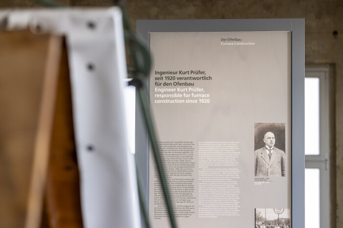 Ausstellungsstele aus der Ferne mit Foto und Text, links Teile eines Zeichenpultes