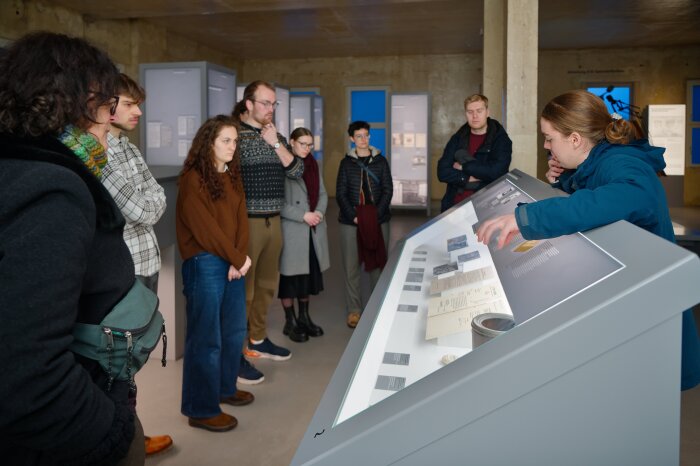 Ein Guide zeigt einer Gruppe Ausstellungsobjekte in einem Pult