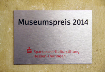 Farbfoto, gelbe Wand, auf der eine Plakette aufgeschraubt ist mit Schriftzug Museumspreis 2014 Sparkassen-Kulturstiftung Hessen-Thüringen