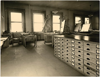 Schwarz-Weiß-Fotografie, rechts Schrank, im Hintergrund Fensterreihe, davor Zeichenmaschinen und Schreibtische, links zwei Waschbecken