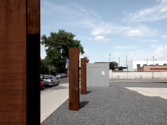 Foto: Spuren im Gelände © Stadtverwaltung Erfurt, Kastner Pichler Architekten Drei Doppelstehlen aus Industriestahl begrenzen eine graue aus Steinen bestehende Fläche. Links im Bild im Straße mit Autos und Gehweg