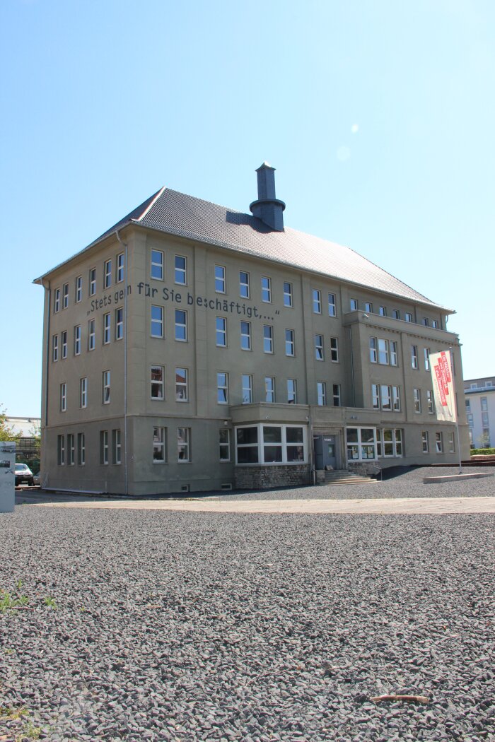 Blick auf den Erinnerungsort Topf & Söhne Ein vierstöckiges Gebäude mit Vorplatz auf dem eine Fahne steht. Der Vorplatz ist mit Schotter gefüllt.