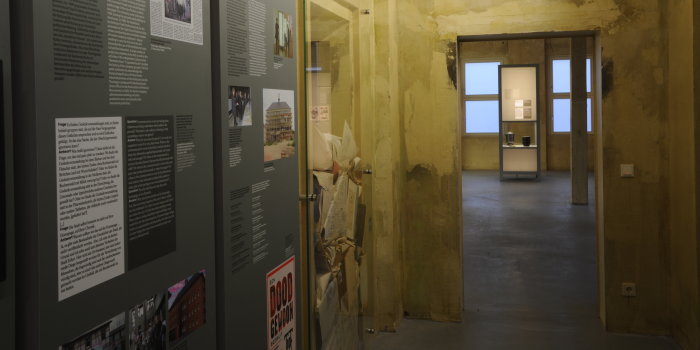 Farbfotografie, links graue Stelen mit Text und Bild, daneben Glasvitrine mit Papier befüllt, rechts im Bild Durchgang mit Blick auf weitere Stele