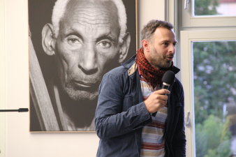 Stehende mittelalterliche männliche Person mit Mikrofon in der Hand, sprichtIm Hintergrund übergroße Fotografie eines alten Mannes