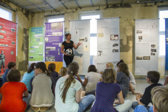 Farbfoto, im Vordergrund sitzt eine Gruppe junger Menschen, im Hintergrund verschiedene bunte Banner, davor eine weibliche Person stehend, die einen Fußball in der Hand hält