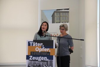 zwei Frauen an einem Mikrofon, eine davon hält eine Rede, davor ein Poster mit der Aufschrift Täter, Opfer, Zeugen 