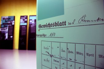 Ausschnitt einer Vitrine im Vordergrund, zu lesen ist u.a. "Gewichtsblatt". Im Hintergrund Ausstellungstafeln. 